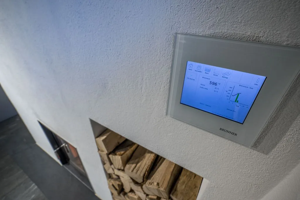 Bedienfeld einer Heizungsregelung an der Wand, darunter ein Fach mit Brennholz